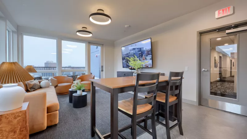 Bright, modern lounge with orange sofas, a wooden high-top table with chairs, large windows, and a wall-mounted TV.