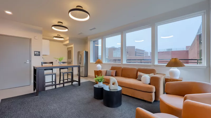 Modern open-plan living room and kitchen with orange furniture, large windows, dark gray rug, and circular ceiling lights.