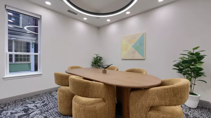 Contemporary meeting room with an oval wooden table, six textured golden-brown chairs, geometric wall art, and plants.