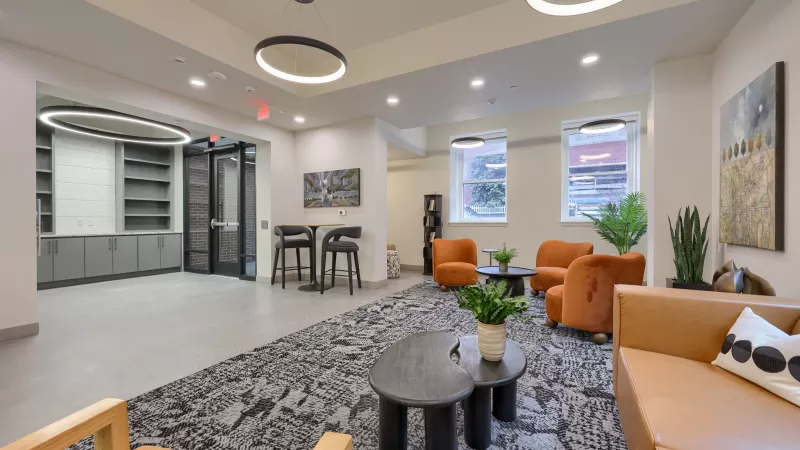 Spacious, modern common lounge with orange armchairs, a brown sofa, high-top seating, and decorative plants under circular lights.