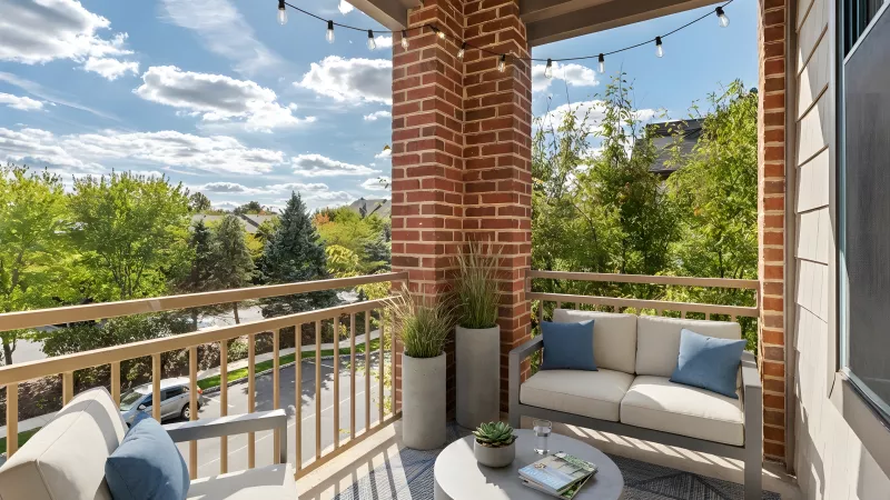 Sunny outdoor balcony with brick, wood railing, a sofa, armchair, coffee table, potted plants, and string lights.