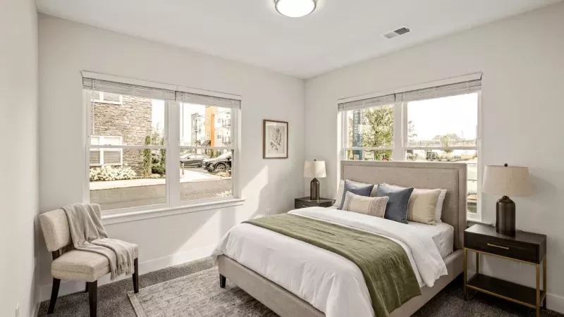 Bright modern bedroom with a bed, white and green bedding, a dark nightstand with lamp, and two large windows.