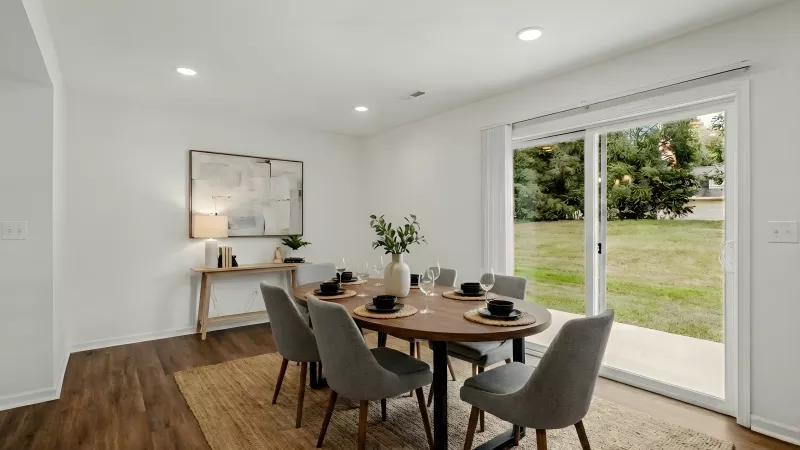 Modern dining room with an oval wood table, six gray chairs, and a sliding glass door to a green backyard.