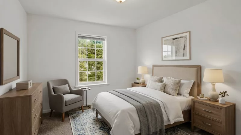 Bright bedroom with a bed, two nightstands and lamps, dresser, mirror, grey armchair, blue rug, and a window with green trees.