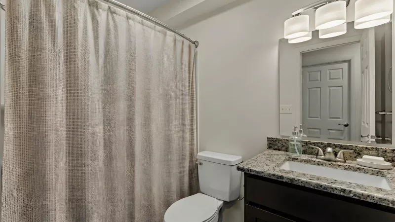 Clean bathroom features a patterned shower curtain, white toilet, dark wood vanity with granite counter, sink, mirror, and lights.
