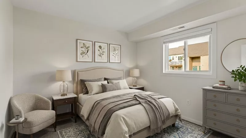 Bright, modern bedroom with a queen bed, two nightstands, lamps, a chair, and a gray dresser. A window overlooks buildings.