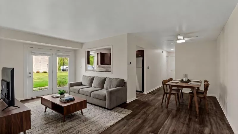 Open-plan living room with grey sofa, coffee table, and TV; dining area with wood table and chairs. Dark floors, glass patio doors.