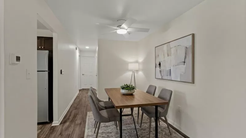 Modern dining room with a wooden table, four grey chairs, a patterned rug, and abstract wall art. Kitchen visible nearby.