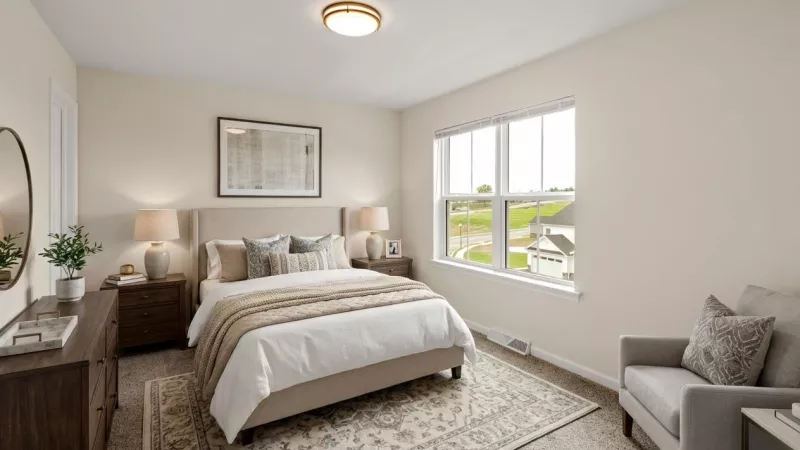 Bright, neutral bedroom with a beige bed, two nightstands, dresser, armchair, and a window looking out onto a neighborhood.