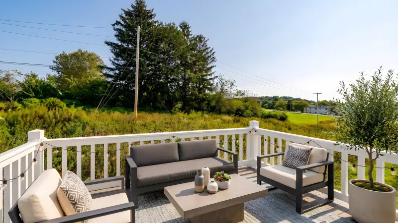 Sunny outdoor deck with modern grey sofa, two beige armchairs, and coffee table on a patterned rug, overlooking a green landscape.