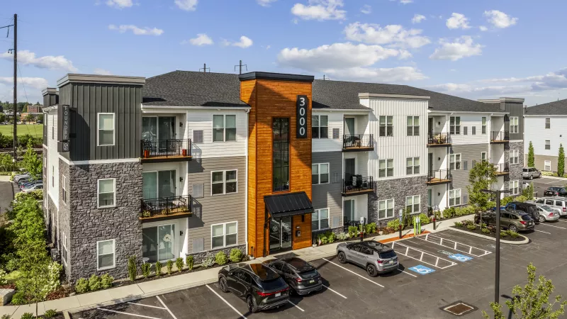 Modern apartment building with dark gray, white, and textured orange-brown facade, multiple balconies, and a parking lot.