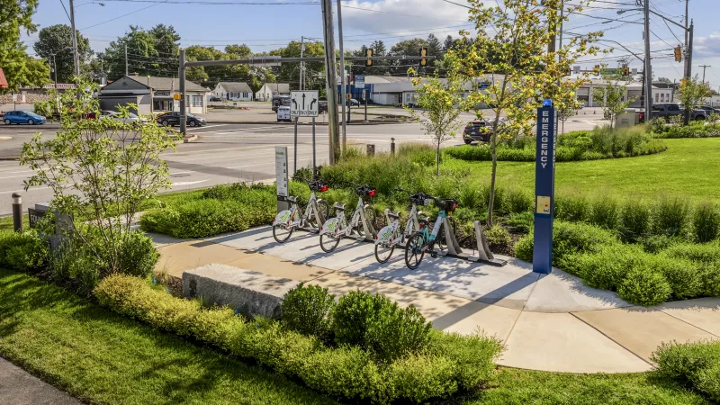 Sunny day at a bike share station with five white and light blue bicycles docked on a concrete pad amidst green landscaping.