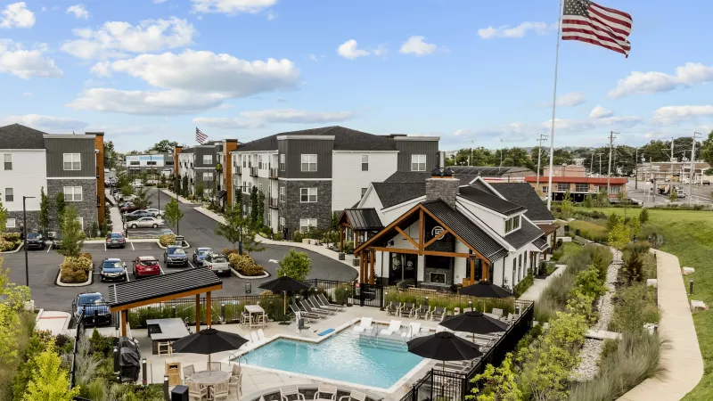 Photograph: Modern apartment complex with a swimming pool, clubhouse, and buildings, under a blue sky. An American flag flies high.