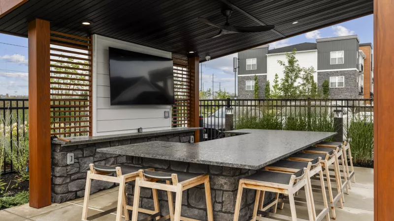 Outdoor covered bar with granite counter, bar stools, and TV. Modern apartment building in the background.