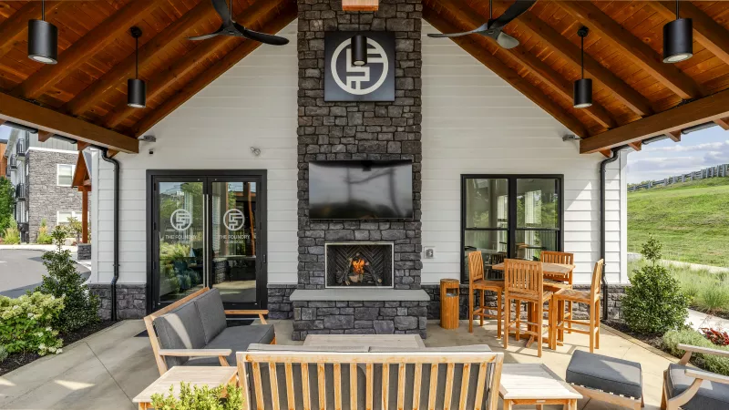 Covered outdoor patio with stone fireplace, TV, and monochrome logo. Furnished with sofas, chairs, and a high-top table.