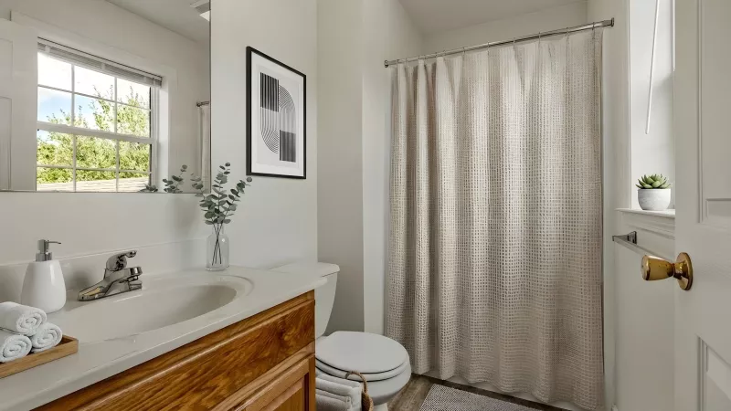 Bright, neutral bathroom with a wooden vanity, white sink, mirror, toilet, and a beige shower curtain.