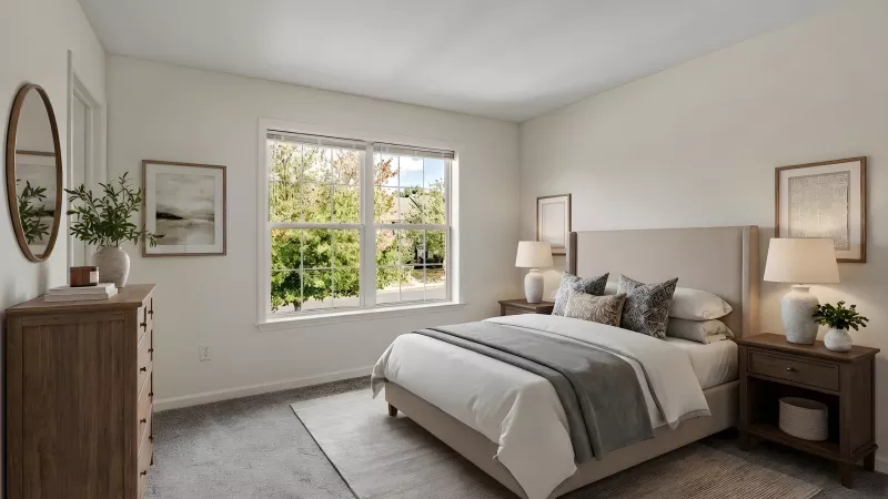 Bright bedroom with a bed, two nightstands, lamps, and a window overlooking trees. A wooden dresser with a mirror is on the left.