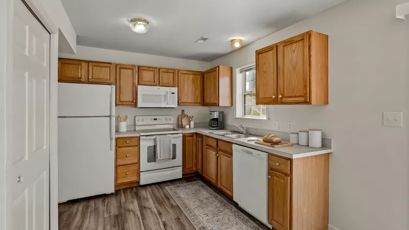 L-shaped kitchen featuring light wood cabinets, white appliances, light gray countertops, and dark wood-look flooring.