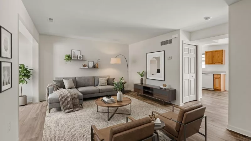 Bright modern living room with a gray sectional, two brown armchairs, coffee table, and TV stand. Kitchen in background.