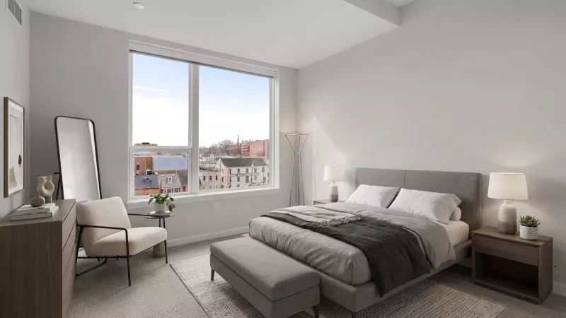 Modern bedroom with a grey bed, two nightstands, a full-length mirror, an armchair, and a large window overlooking city buildings.