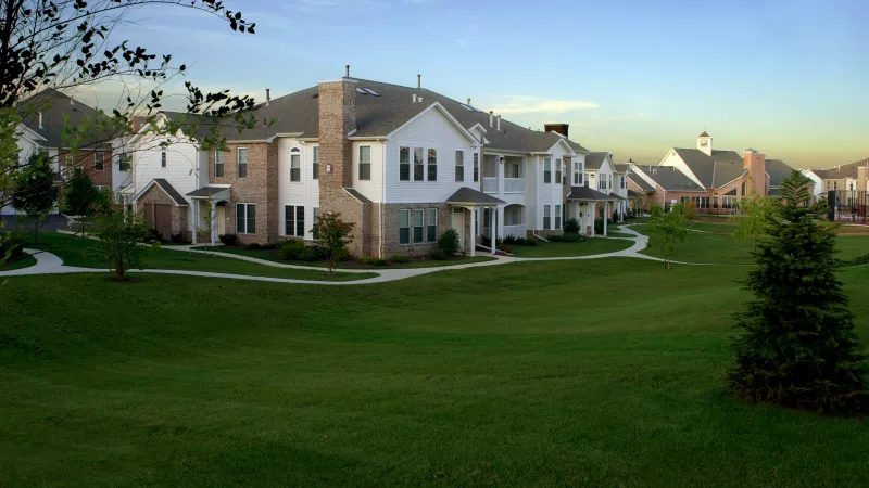 Row of modern townhouses with white siding and brick accents, facing a large, well-maintained green lawn under a blue sky.