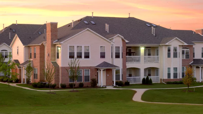 Apartment building with beige siding and brick accents, green lawn, and a beautiful orange and pink sunset sky.