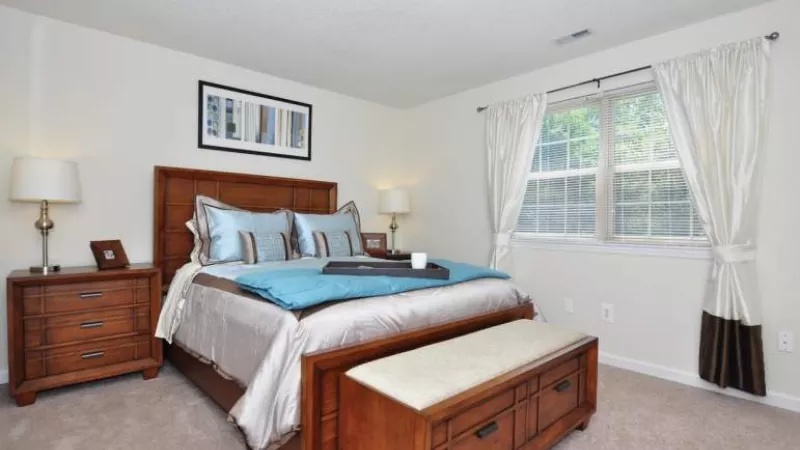 Cozy bedroom with a wooden bed, blue and brown bedding, nightstands, lamps, a window with white curtains, and a storage bench.