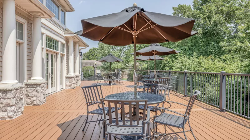 Outdoor wooden deck with black patio furniture and umbrellas. A light-colored building and green trees are visible.