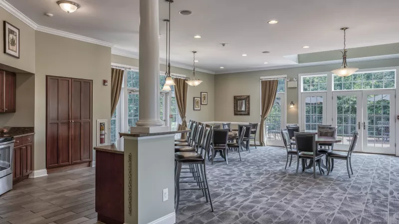 Spacious community room with a kitchen, bar stools, dining tables, patterned carpet, and large windows.