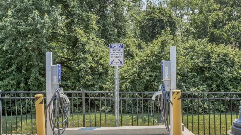 Outdoor EV charging station with two chargers, a sign, yellow bollards, a black fence, and green trees in the background.
