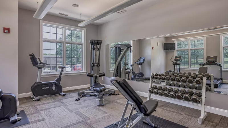 Photograph of a bright fitness center with exercise bikes, a weight bench, and a large rack of dumbbells, reflecting in a wall mirror.