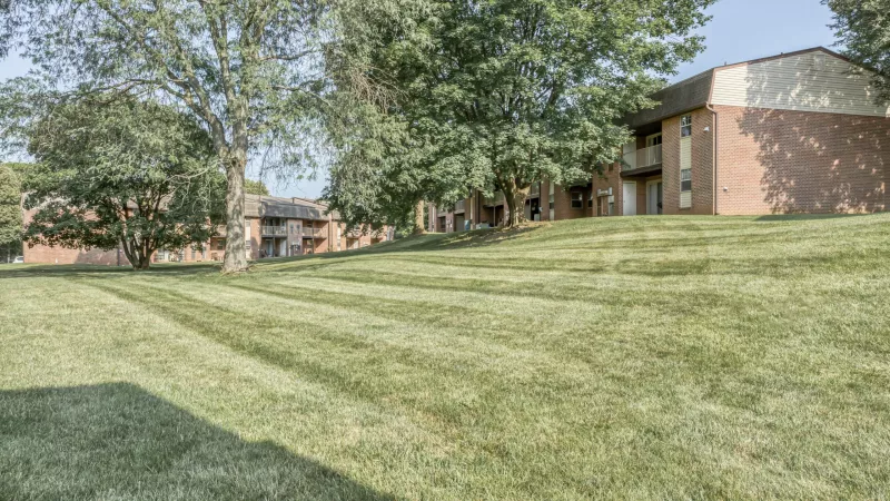 Expansive green lawn sloping towards brick apartment buildings under a clear sky, with several large trees.