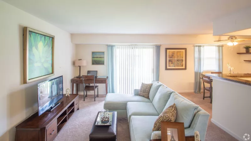 Bright living room features a light blue sectional sofa, TV, desk, and adjoining dining space, illuminated by a sliding glass door.