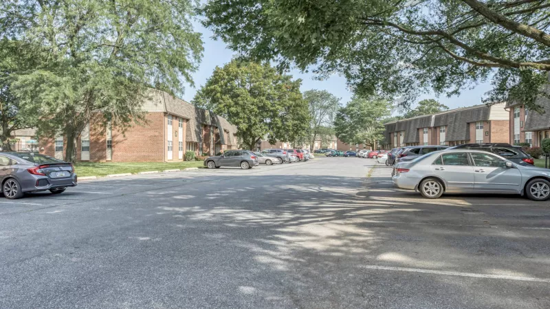 Outdoor parking lot with cars, multi-story apartment buildings, and green trees under a clear sky.