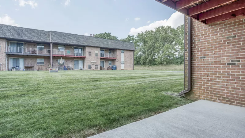 View from a patio of a large grassy lawn leading to two-story apartment buildings with balconies. Brick wall on right.