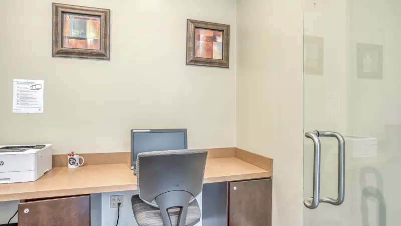 A small business center with a computer on a light brown desk, printer, and office chair. Two abstract paintings hang on the wall.