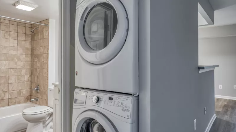 White stacked washer and dryer. Tiled bathroom with toilet and tub to the left, gray room with dark floor to the right.