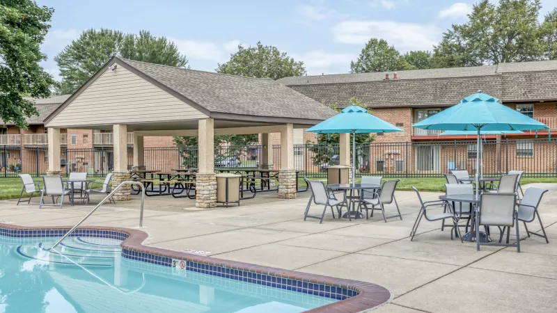 Apartment complex pool area with patio tables, two blue umbrellas, and a large covered pavilion.