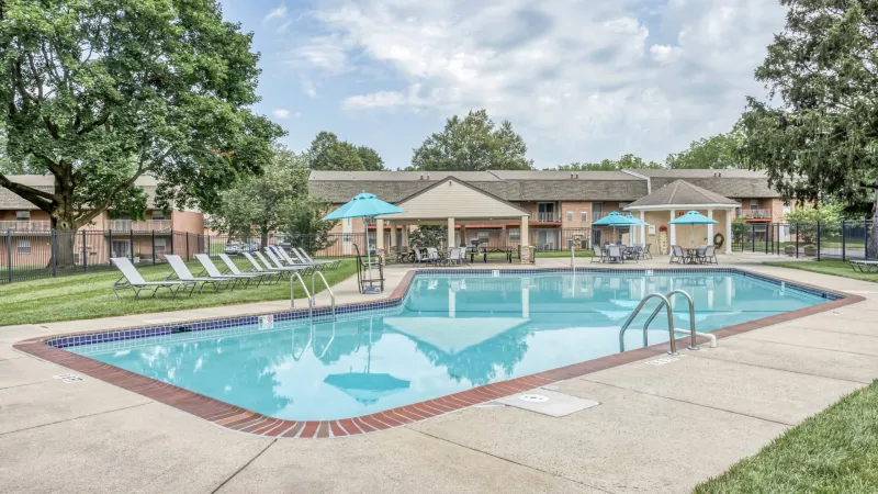 Outdoor swimming pool with lounge chairs, covered patio, blue umbrellas, and apartment buildings under a partly cloudy sky.