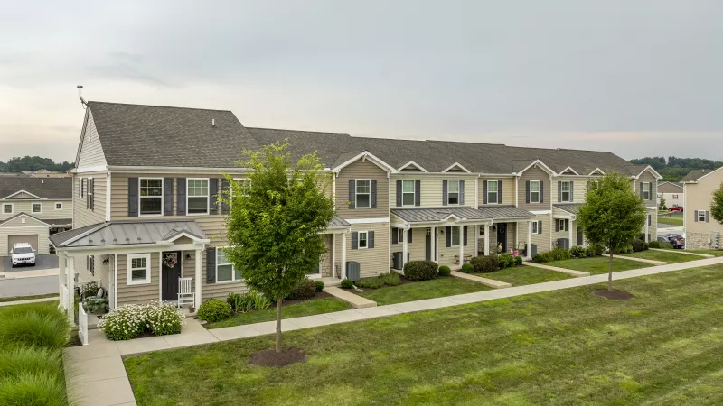 Row of modern townhouses with light siding, dark roofs, and small trees on a green lawn with a sidewalk.