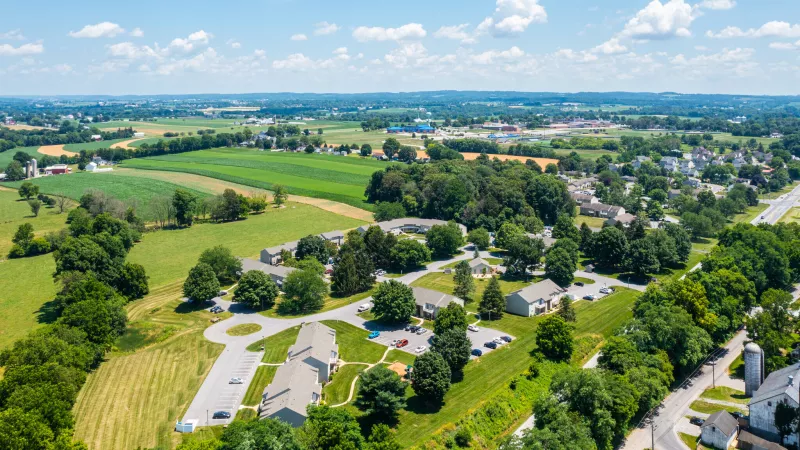 A scenic drone image showcasing the Pioneer Woods apartment community in Lancaster, PA, nestled among open green fields and rolling farmland under a blue summer sky.