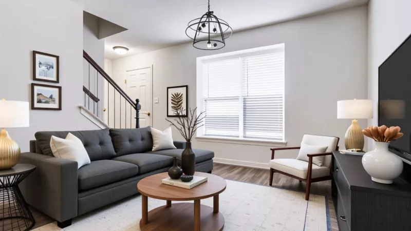 Modern living room at Mill Creek Manor Apartments with a gray sofa and white pillows, round wood coffee table on a light area rug, accent chair, media console with lamp and vase, large window with blinds, staircase, and contemporary pendant light.