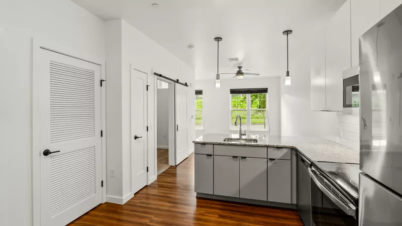 Modern apartment kitchen at Stadium Row featuring stainless steel appliances, granite countertops, sleek white cabinetry, and wood-style flooring with natural light from adjacent living area