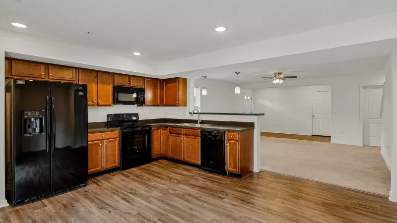 A view of the fully-equipped kitchen in a Parker townhome, showcasing dark wood cabinetry, black appliances, including a large refrigerator, microwave, and dishwasher, and ample counter space with a breakfast bar that opens to the living area.