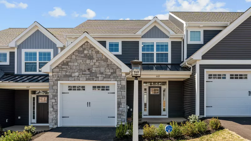 Front exterior view of modern townhomes with dark siding, white trim, stone accents, and two-car garages. A landscaped sidewalk leads to the front door of the center unit.