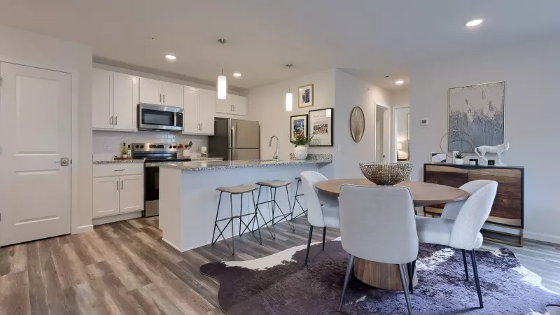 Open kitchen at The Pond Apartments with white cabinetry, stainless steel appliances, granite countertops, and a dining area with round table and upholstered chairs.