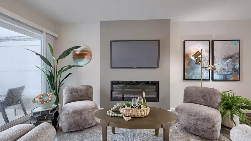 Cozy living room at The Pond Apartments with modern fireplace, wall-mounted TV, two armchairs, coffee table, and large decorative plants.