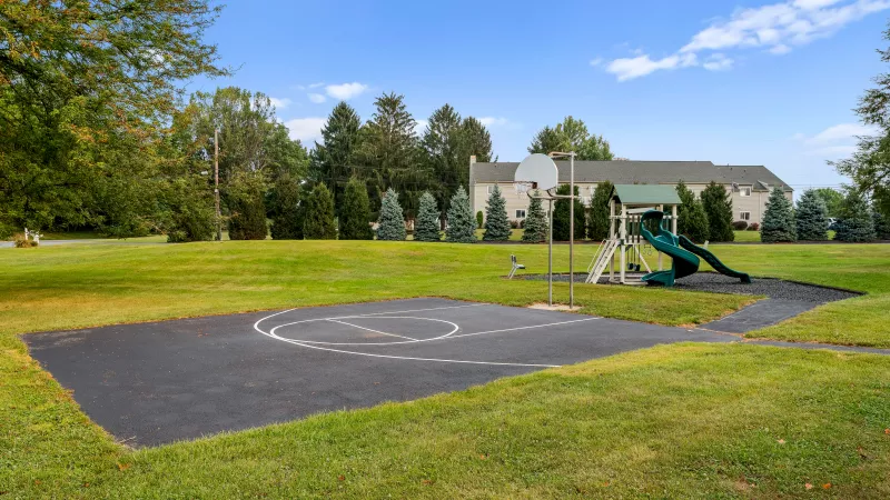 Outdoor basketball half-court and playground with slide, set in a grassy open area with trees and apartments in the background.