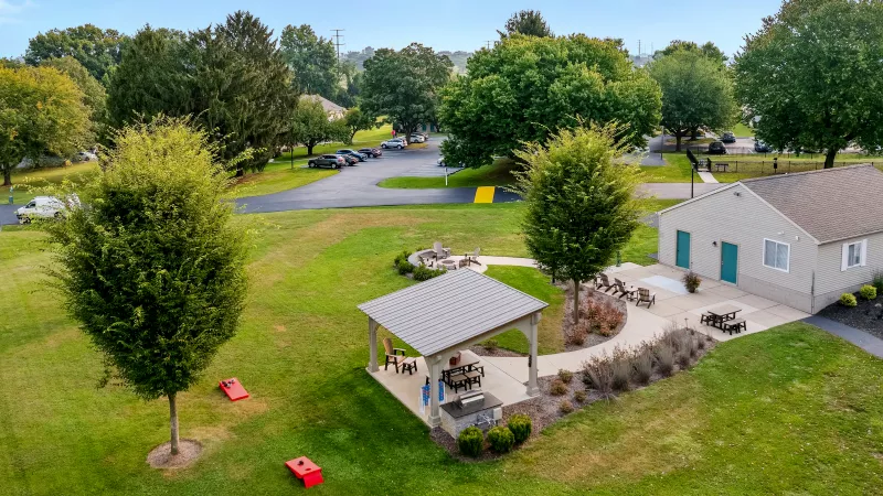Aerial view of community green space with covered picnic pavilion, outdoor seating, fire pit, and cornhole boards surrounded by trees and parking areas.