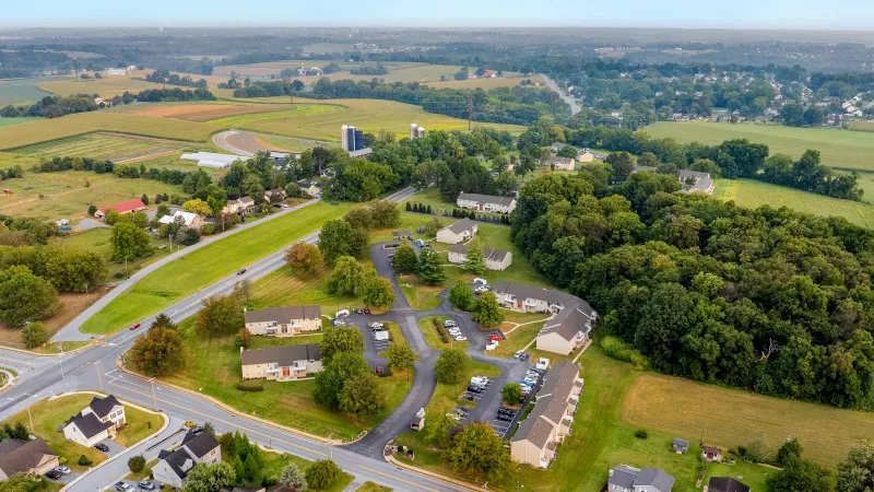Aerial view of the Pioneer Woods apartment community surrounded by farmland, roads, and trees with parking lots and townhome-style buildings visible.